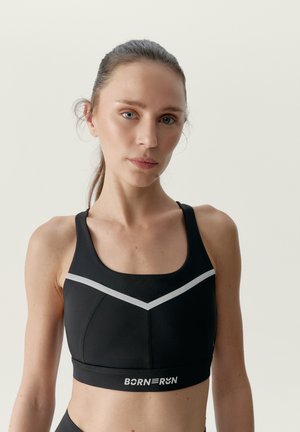 Frau mit heller Haut und zurückgebundenem Haar, die einen schwarzen Sport-BH mit weißen Streifen und dem Text "BORN RUN" auf dem Bund trägt.