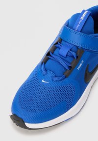 Baskets Nike bleues avec matériau en mesh, sangle de maintien dynamique, accents noirs, surface texturée et semelle blanche. Logo swoosh noir bien visible.