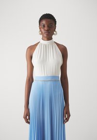 Haut à col halter en tissu blanc cassé associé à une jupe fluide plissée de couleur bleu clair ; présente une ceinture ornée de bijoux.