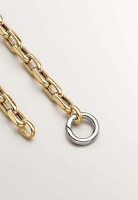 Cadena de oro con eslabones alargados de un lado, que transiciona a un gran anillo circular de plata. Textura suave con un brillo metálico.