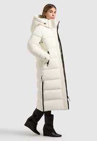 Abrigo puff largo blanco con capucha, que cuenta con bolsillos con cremallera y detalles en negro. Combinado con botas de tobillo negras con cuña.