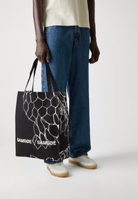 Borsa tote nera con grafica floreale bianca e branding. Realizzata in tessuto, presenta doppi manici e una forma rettangolare.