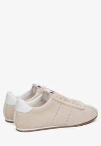 Beżowe zamszowe sneakersy z białymi akcentami, z przeszyciami, płaskimi sznurowadłami i gumową podeszwą. Widok z tyłu pokazujący zakrzywiony design pięty.