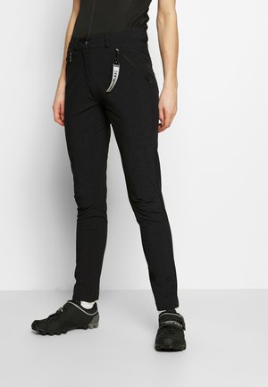 Person wearing fitted black pants with zipper pockets and black cycling shoes with white socks, standing against a plain background.