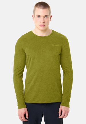 Junger Mann mit kurzen Haaren, der ein olivgrünes Langarmhemd und dunkle Hosen trägt, vor einem schlichten hellen Hintergrund steht.