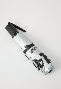 Kompakt ezüst esernyő fekete geometriai mintával, fekete fogantyúval és pánttal. Tartalmazza a "KARL LAGER JEANS" logót.