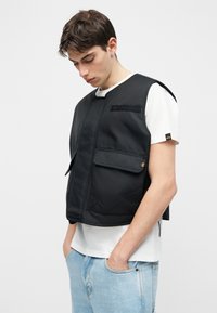 Νέος άνδρας που φοράει μαύρο αμάνικο γιλέκο πάνω από λευκό μπλουζάκι και ανοιχτόχρωμο μπλε τζιν, κοιτάζοντας προς τα κάτω με τα χέρια στις τσέπες.