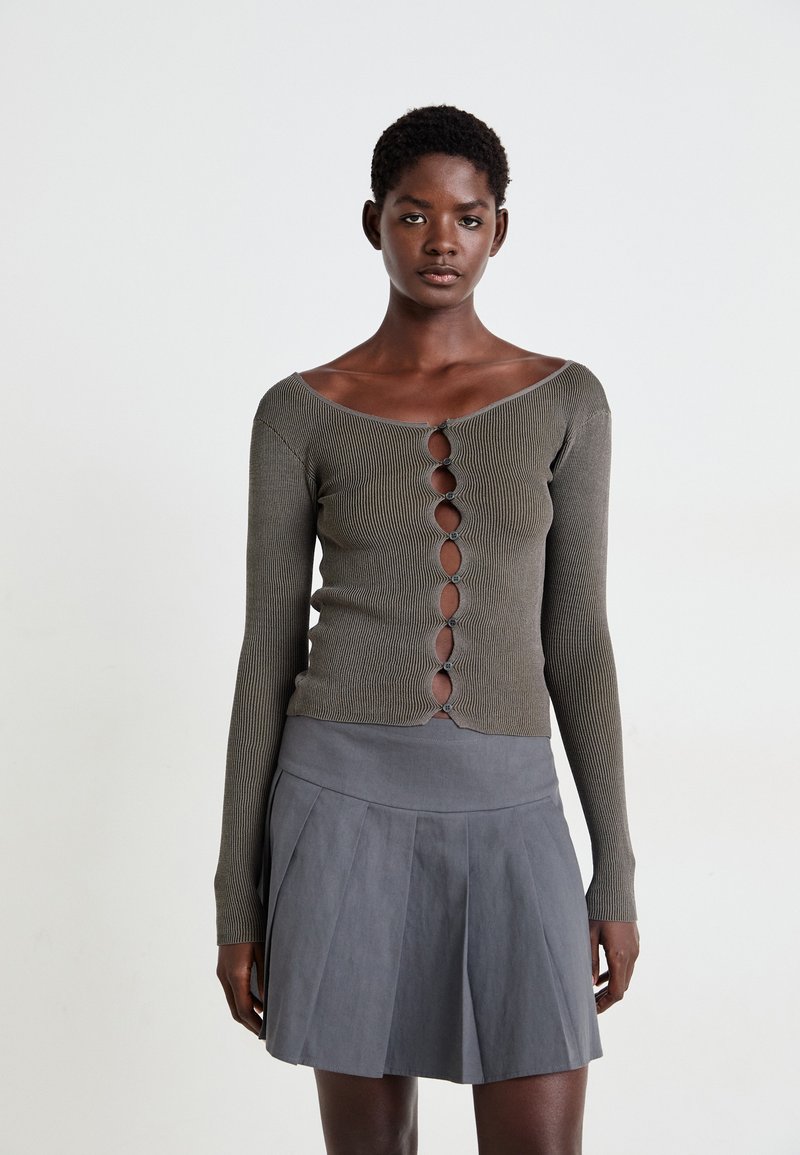 Olijfgroene geribbelde top met lange mouwen, zes knoopsluitingen en off-shoulder ontwerp, gecombineerd met een uitlopende grijze plooienrok.