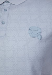 Polo azul claro con un patrón tejido texturizado, que presenta un parche de goma con la forma de una cara estilizada con gafas de sol.