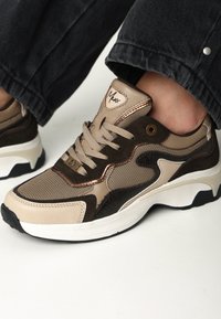 Zapatilla beige y marrón oscuro con materiales de malla y cuero, que presenta cordones redondos, una lengüeta acolchada y una suela blanca y texturizada.