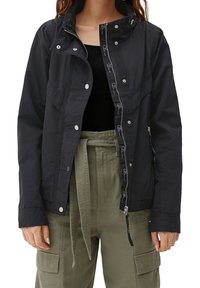 Veste légère noire avec fermetures à pression et à glissière, dotée d'un col montant et de plusieurs poches. Portée sur un haut noir et un pantalon olive.