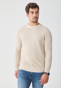 Béžový žebrovaný svetr s kulatým výstřihem a dlouhými rukávy, v kombinaci s modrými džíny. Texturovaný pletený vzor a žebrované manžety a lem. Hnědé hodinky na zápěstí.