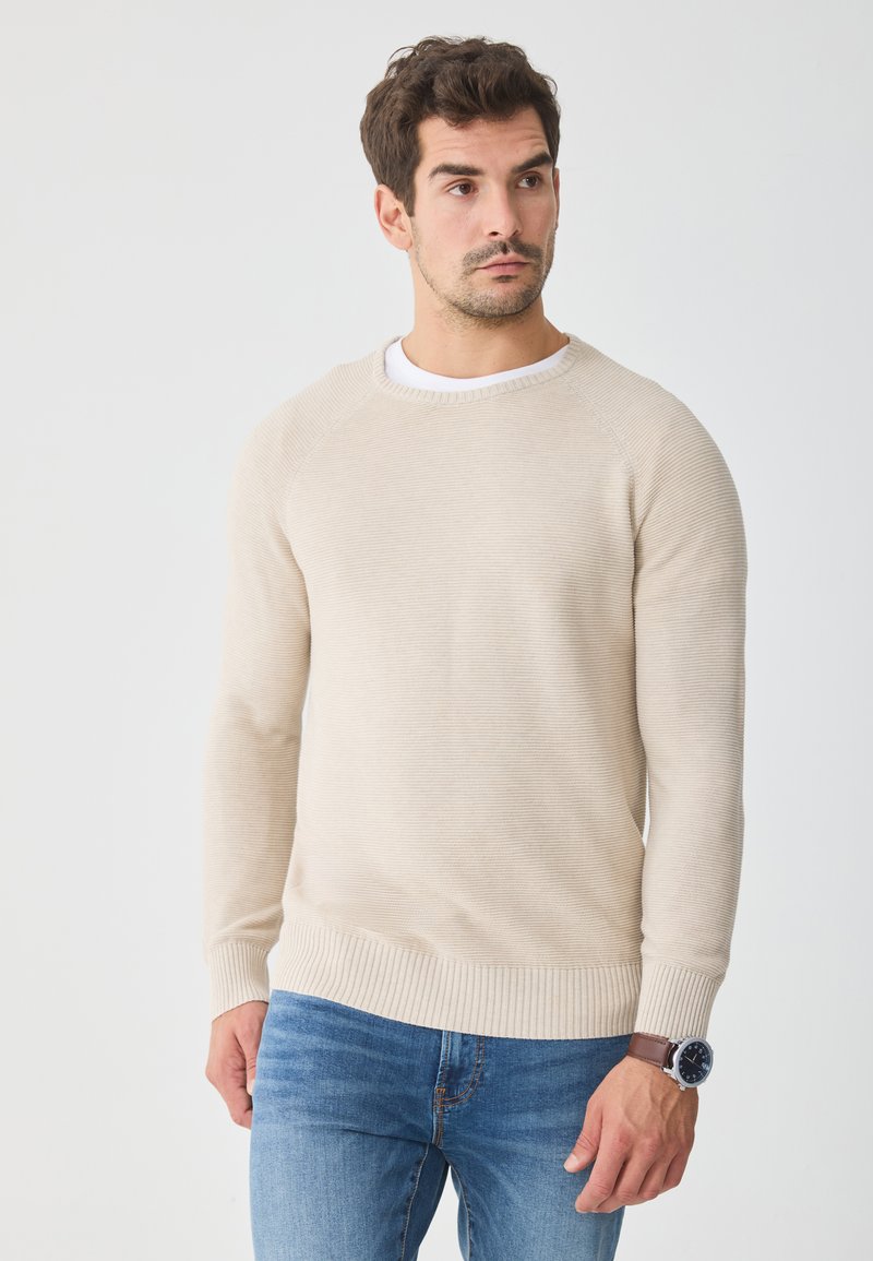 Béžový žebrovaný svetr s kulatým výstřihem a dlouhými rukávy, v kombinaci s modrými džíny. Texturovaný pletený vzor a žebrované manžety a lem. Hnědé hodinky na zápěstí.