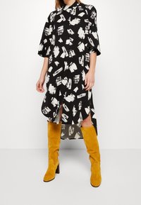 Czarna sukienka koszulowa z białym abstrakcyjnym wzorem, krótkimi rękawami i rozcięciami po bokach. W połączeniu z wysokimi, żółtymi zamszowymi botkami i guzikiem.