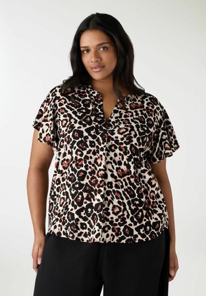 Donna con pelle medio-scura e lunghi capelli scuri, indossa una camicetta a maniche corte con stampa leopardata e pantaloni neri, in piedi davanti a uno sfondo semplice.