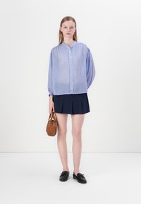 Sininen ja valkoinen raidallinen paita, jossa on napitus edessä, ylisuuri istuvuus ja rypytetyt hihat, yhdistettynä tummansiniseen taitetun hameeseen ja ruskeaan käsilaukkuun.