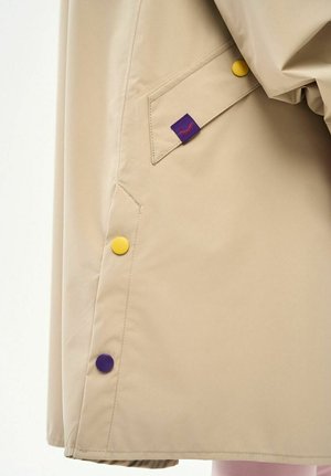 Beżowy rękaw płaszcza z ukośną klapką kieszeni, fioletowo-żółtymi zatrzaskami oraz małą fioletową metką z tkaniny z czerwonym wzorem rzęs.