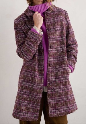 Femme portant un manteau boutonné à carreaux marron et violet sur un pull à col roulé violet vif et un pantalon marron, main près du menton.