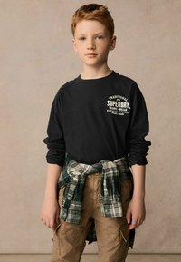 Tricou negru cu mâneci lungi și logo alb, purtat cu pantaloni cargo kaki. O cămașă din flanel cu carouri verzi legată în jurul taliei. Design simplu și casual.