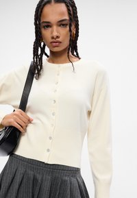 Cardigã creme com bainha canelada e fecho de botão. Usado sobre uma saia escura com riscas finas, apresenta mangas longas e um decote redondo.