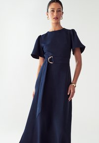 Námornícky modré šaty z hladkej látky, s krátkymi puffovacími rukávmi, priliehavým pásom so zlatou sponou a splývavou sukňou.