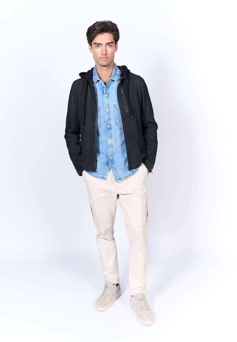 Veste à capuche noire sur une chemise en denim clair, assortie à un pantalon cargo beige et des baskets grises. Textures lisses et couleurs contrastées présentes.