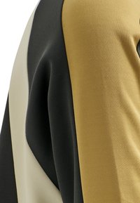 Gros plan sur un tissu avec des blocs de couleur noir, beige et jaune moutarde, montrant les détails de la texture et des coutures.