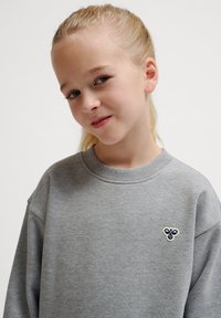 Grå sweatshirt med ribbad halsringning och ärmslut, med en liten broderad logotyp på bröstet. Mjuk textur och avslappnad passform.