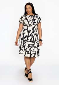 Schwarzes und cremefarbenes Kleid mit kurzen Ärmeln, tailliertem Gürtel und abstraktem Blumenmuster; kombiniert mit schwarzen Espadrilles.