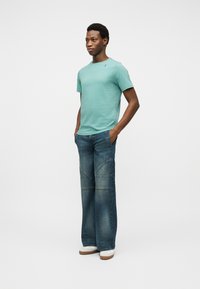 Άνδρας στέκετα ι με τα χέρια στις τσέπες, φοράει μπλε-πράσινο κοντομάνικο πουκάμισο, φαρδιά μπλε τζιν και λευκά αθλητικά παπούτσια μπροστά σε απλό φόντο.