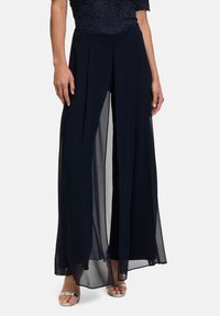Granatowe spodnie palazzo wykonane z lekkiego szyfonu, o szerokim kroju nogawki, z plisowanym wzornictwem i przejrzystą warstwą. Widziane srebrne sandały.