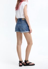 Denim shorts with rolled cuffs, light blue wash, back pockets, paired with a white short-sleeve top and black flat sandals.