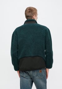 Teal-värinen fleece-takki, jossa on teksturoitu pinta, musta alaosa ja korkea kaulus. Takakuvassa näkyy löysä istuvuus ja rento muotoilu.