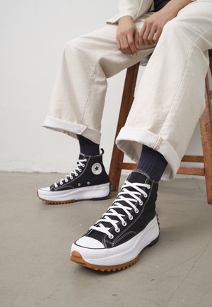 Person siddende iført høje sorte og hvide sneakers med tykke gummisåler, beige opkørte bukser og mørke sokker på grå gulv.