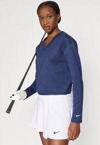 Μπλε ναυτικό μπλουζάκι με μακριά μανίκια και V-λαιμόκοψη συνδυασμένο με λευκή φούστα με πιέτες. Διαθέτει λογότυπο Nike τόσο στο μπλουζάκι όσο και στη φούστα. Κρατά ένα γκολφ κλαμπ με γάντι.