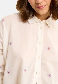 Camicia a maniche lunghe di colore chiaro, realizzata in tessuto morbido, con ricami floreali in blu e arancione. Colletto classico e texture sottile.