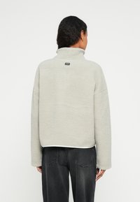 Jasnoszara bluza polarowa z wysokim kołnierzem, opuszczonymi ramionami oraz małą naszywką z logo na plecach. Wykonana z miękkiego, teksturowanego materiału.