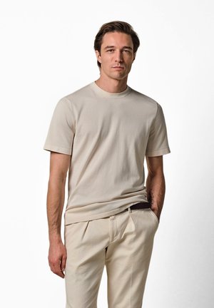Mann trägt beige kurzärmeliges T-Shirt und passende Hose, steht mit einer Hand in der Tasche vor weißem Hintergrund.