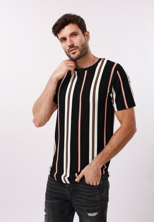 Homme aux cheveux bruns courts et à la barbe, portant un t-shirt noir avec des rayures verticales blanches et marron, posant avec une main près du menton et l'autre dans la poche.