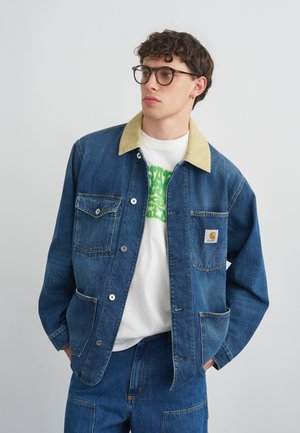 Joven con pelo rizado con gafas, chaqueta vaquera azul con cuello beige y pantalones vaqueros azules a juego, manos en los bolsillos.