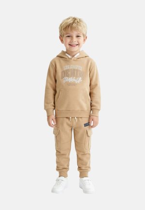 Jeune garçon aux cheveux blonds portant un sweat à capuche beige Lee Cooper et un pantalon cargo assorti, souriant et posant devant un fond blanc.