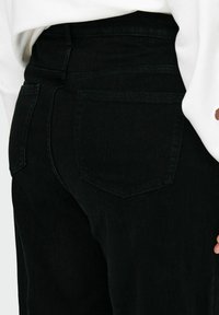 Jean en denim noir à taille haute, avec deux poches arrière et une texture lisse, porté avec un haut blanc partiellement visible.