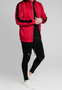 Chaqueta roja con cremallera y rayas negras en los laterales, llevada sobre una camiseta negra. Combinada con jeans negros desgastados y zapatillas blancas.