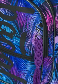 Zaino con un motivo di foglie tropicali blu e viola, accenti neri, una cerniera con clip metallica e un dettaglio di frangia in macramè viola.