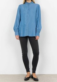 Camicia in denim azzurro chiaro con collo alto, maniche lunghe a sbuffo e chiusura con bottoni, abbinata a pantaloni neri aderenti e scarpe flat nere.