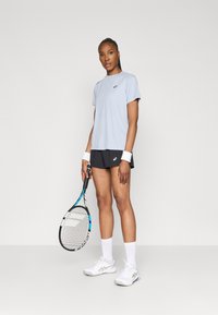 Ανοιχτό μπλε κοντομάνικο αθλητικό μπλουζάκι συνδυασμένο με μαύρες βερμούδες. Λευκά παπούτσια τένις και φίμους. Κρατάει μια ρακέτα τένις.