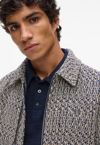 Kravejakke med krave og lynlås, i et tekstureret, vævet mønster i blå, creme og gyldne nuancer. Har et lille logo på brystet og en mørk polo under.