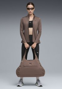 Brun sportstaske med en bred rem, lynlåsrum og glat overflade, båret af en person i atletisk tøj mod en gradient baggrund.