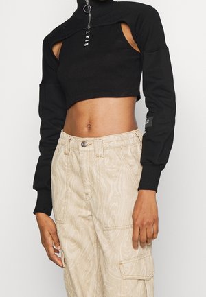 Person trägt ein schwarzes, kurz geschnittenes Langarmshirt mit Aussparungen an den Schultern und beige, hoch taillierte Cargo-Hosen mit Fronttaschen.