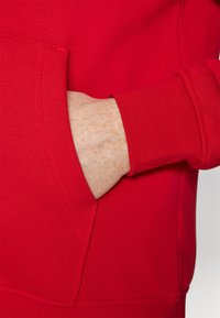 Hand met sproeten gedeeltelijk in de voorzak van een rode lange mouwen sweatshirt met geribbelde manchetten.